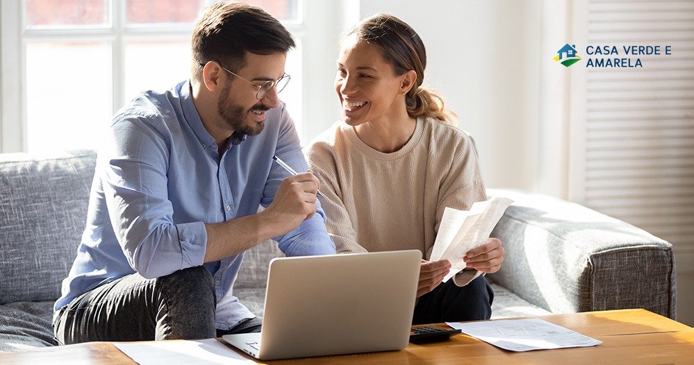 Casal sorridente no sofá analisa documentos e um notebook, com o selo do programa Casa Verde e Amarela no canto