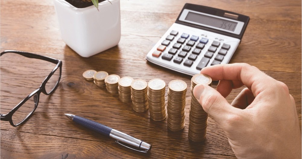 Mão empilha moedas em colunas crescentes sobre uma mesa de madeira, ao lado de calculadora, óculos e caneta