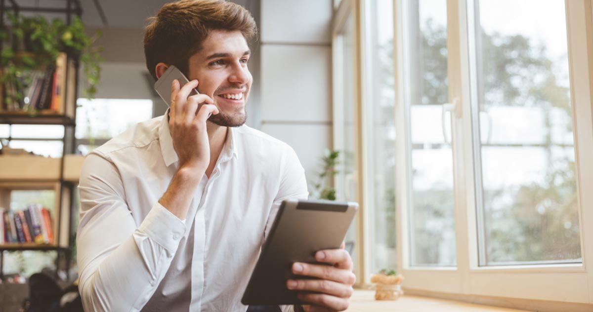 Homem sorridente fala ao celular segurando um tablet, em um escritório claro