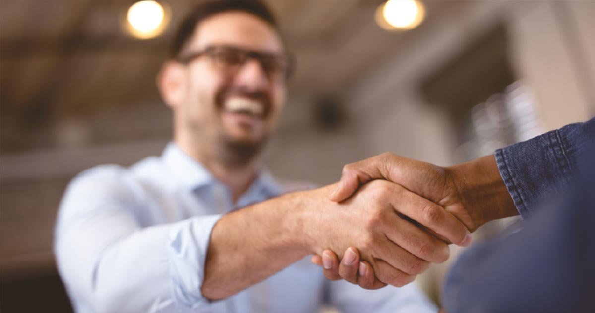 Aperto de mãos entre um homem sorridente de óculos e outra pessoa, fechando negócio