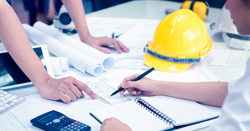 Profissionais analisam plantas de arquitetura, com calculadora e capacete amarelo sobre a mesa