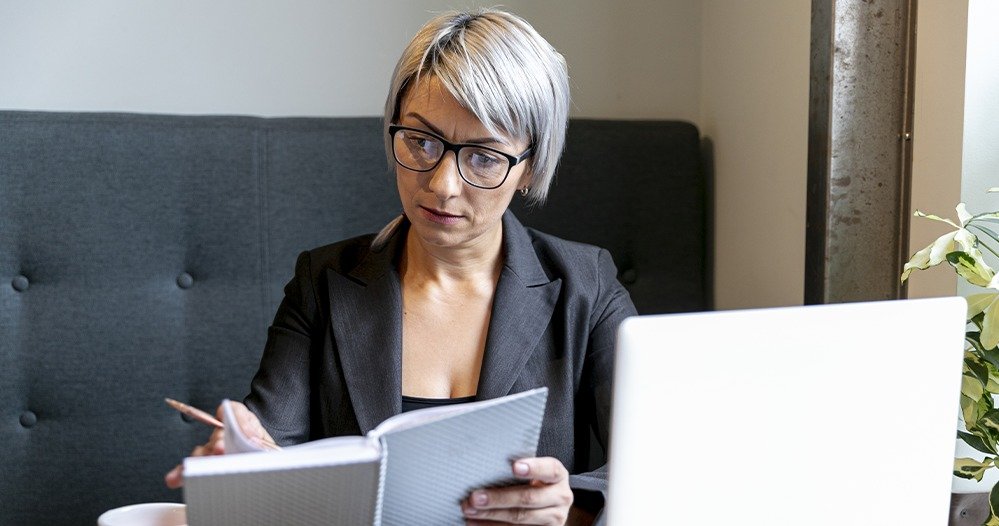 Mulher de óculos lê um caderno de anotações em frente a um notebook